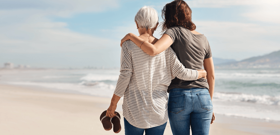 A young woman and an elderly woman standing together on a beach, looking at the ocean. The elderly woman has short grey hair and is wearing a striped shirt, while the young woman is in a gray t-shirt and blue jeans. They are embracing each other, sharing a moment of connection.